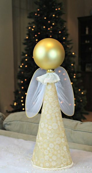 Un ángel creado con un cono y una bola dorada con un árbol de navidad al fondo.